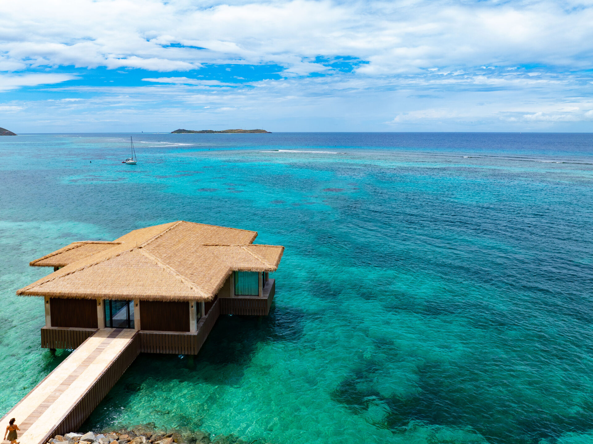 A thatched-roof overwater bungalow sits on turquoise, clear ocean water at Oil Nut Bay in the British Virgin Islands, with a wooden walkway leading to it, surrounded by coral reefs and distant islands under a partly cloudy sky.