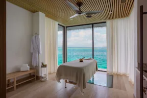 A serene spa room at Oil Nut Bay with a massage table facing large windows overlooking the turquoise ocean. White curtains, a bathrobe on the wall, and wooden furnishings create a tranquil, relaxing British Virgin Islands atmosphere.