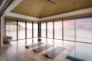 A serene yoga studio at Oil Nut Bay’s Overwater Spa, with three mats and yoga blocks, faces floor-to-ceiling windows overlooking a calm ocean and colorful sunset sky in the British Virgin Islands, complete with a modern chair and gong in the corner.
