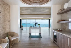 A modern, coastal living space with wicker chairs, stone walls, and an open view of the ocean through large glass doors. Inspired by Oil Nut Bay spa, minimalist decor and a stylish ceiling accent create a serene, airy atmosphere.
