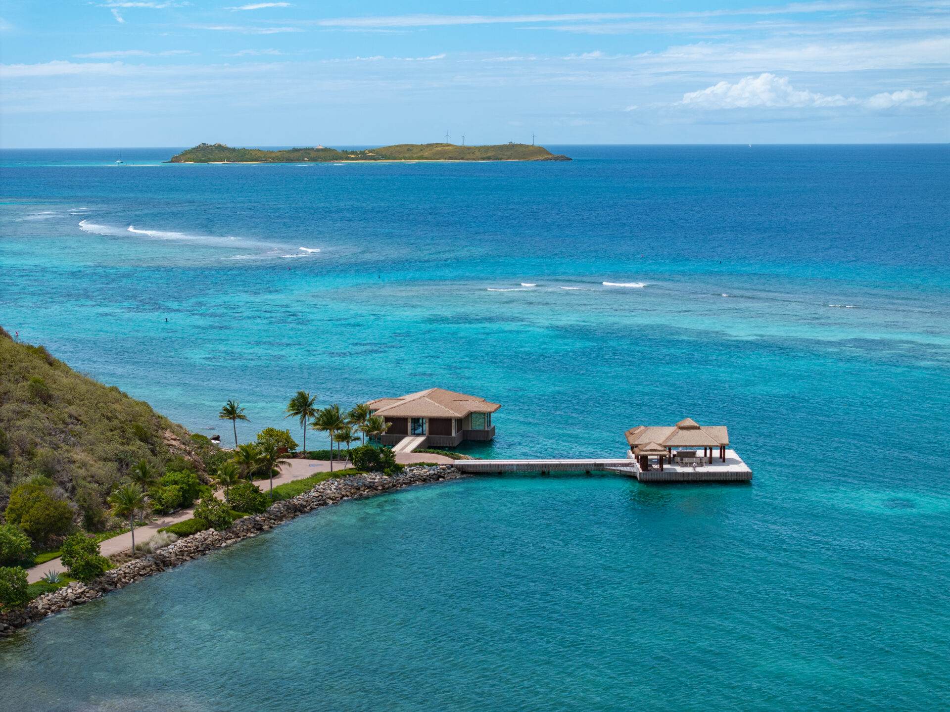 Aerial view of a tropical coastline featuring the Oil Nut Bay spa, with a dock and two buildings over clear turquoise water, surrounded by lush greenery and palm trees, and a small island in the distance under a blue sky.
