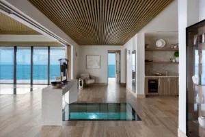 Modern open-plan room with wood flooring, glass floor panel revealing water below, floor-to-ceiling windows with an ocean view—perfect for relaxing after a day at the Oil Nut Bay spa. Includes kitchenette, shelves, and a cozy armchair by the far door.