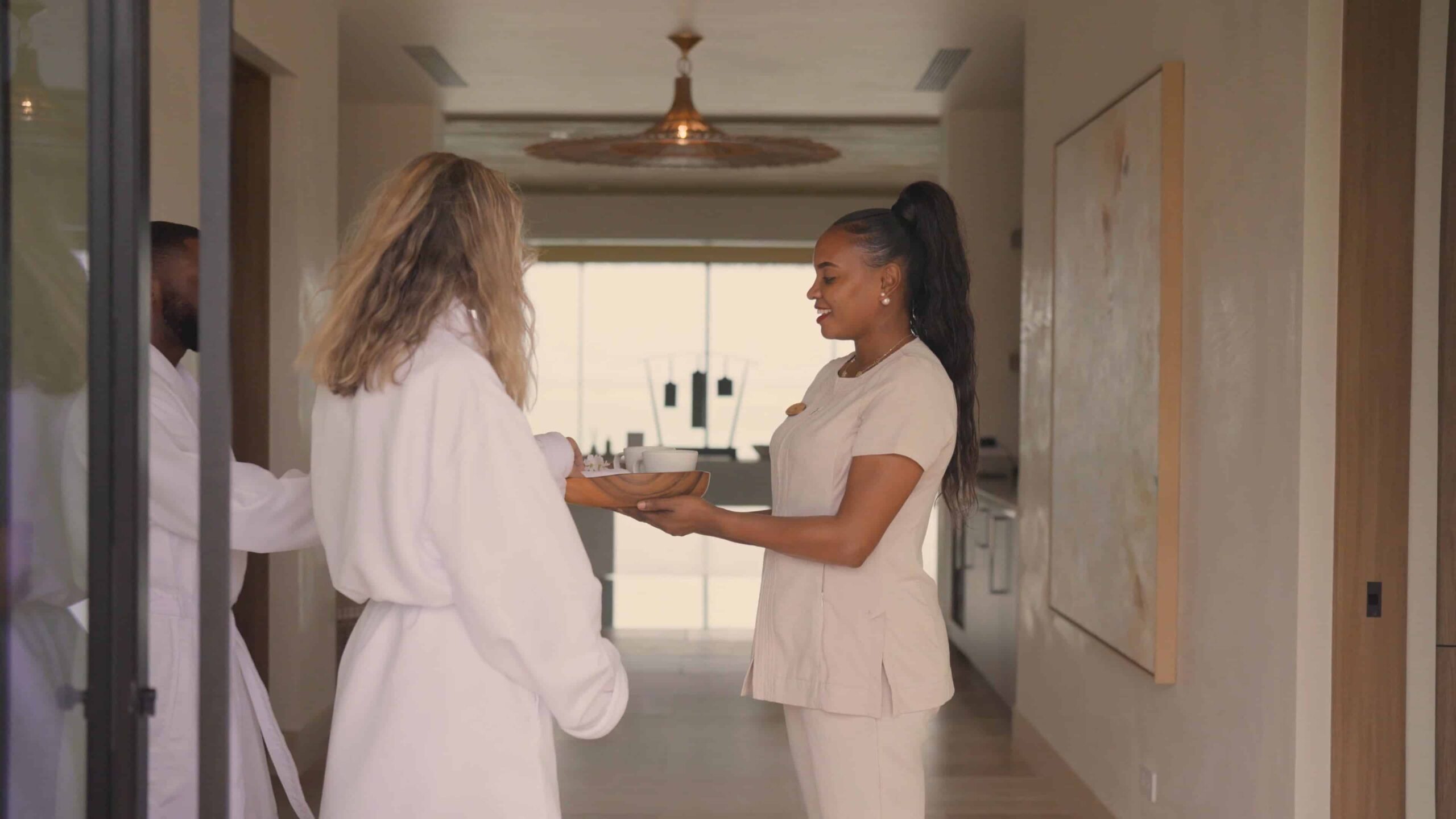 A smiling woman in beige spa attire hands a wooden tray with drinks to a person in a white robe in Sundara Spa’s bright, modern hallway adorned with large art and natural light.