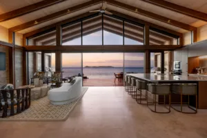 Spacious, modern open-concept living room and kitchen with vaulted wooden ceilings, large windows, and sunset ocean views. Enjoy the outdoor patio and islands through glass doors in these new villas at Oil Nut Bay, British Virgin Islands.