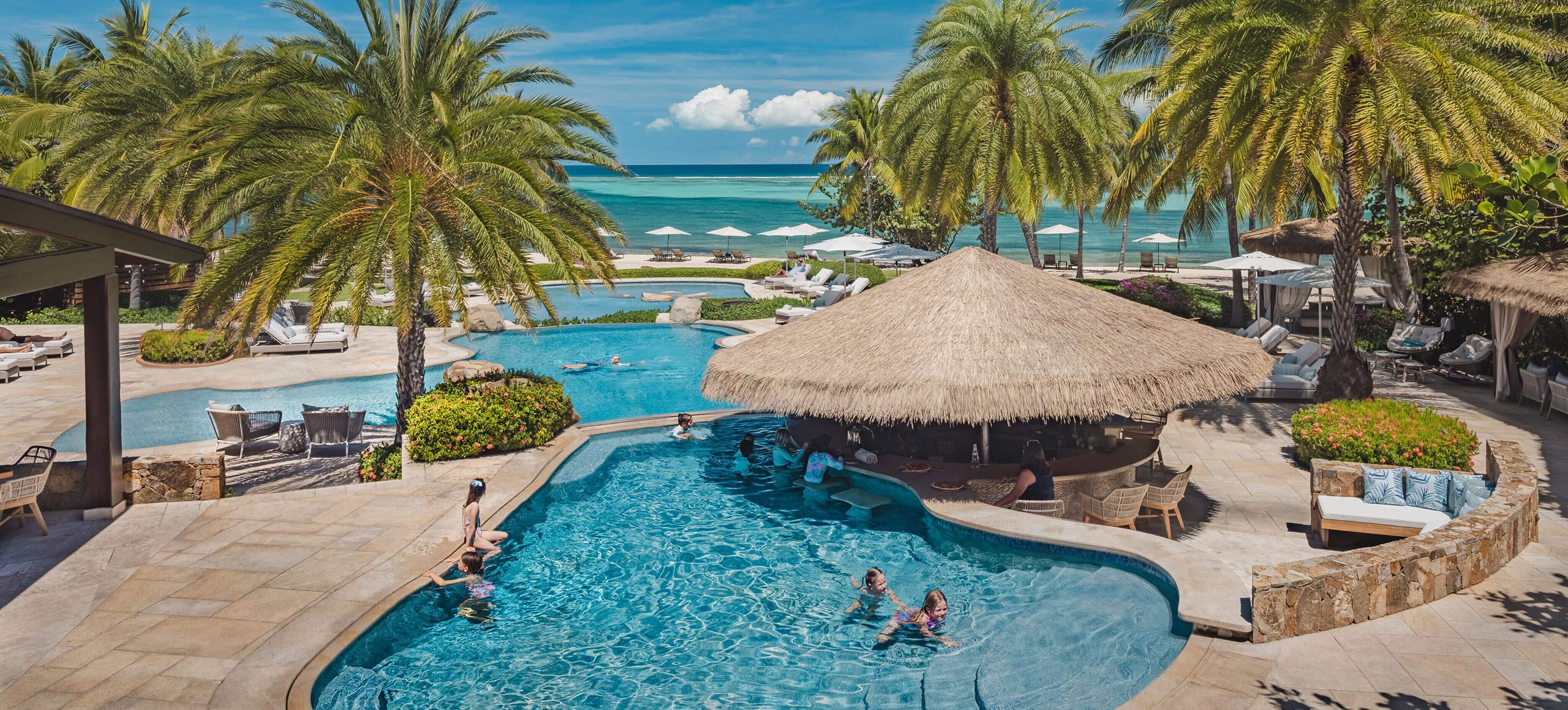 A tropical resort in Oil Nut Bay features multiple pools surrounded by palm trees. One pool has a thatched-roof bar with people sitting on bar stools in the water. Lounge chairs and umbrellas dot the area, while the ocean and a clear blue sky are visible in the background.