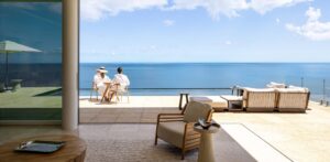 Two people in white robes sit at a table on a spacious patio with a sea view in Oil Nut Bay. Modern furniture, including chairs and a sofa, is placed around the area. A large glass window is on the left, and the clear blue sky meets the ocean horizon in the background.