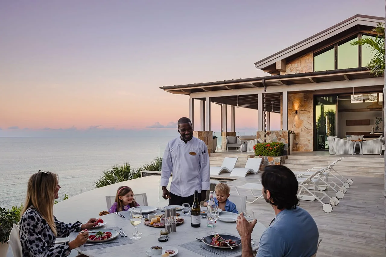 A family of four dines outdoors with a view of the ocean at sunset in Oil Nut Bay. A waiter stands beside the table, engaging with them. The table is set with plates of food, glasses of wine, and a bottle of wine. The modern house in the background has large windows and outdoor seating.