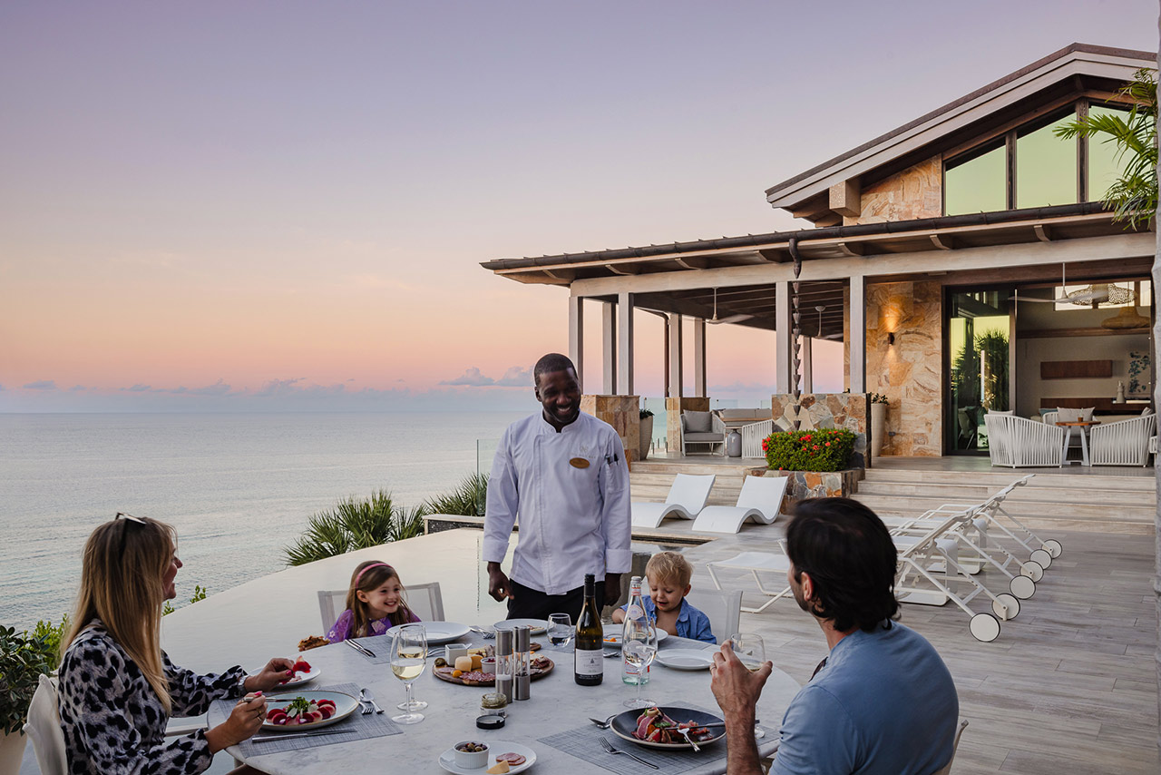 A family of four dines outdoors with a view of the ocean at sunset in Oil Nut Bay. A waiter stands beside the table, engaging with them. The table is set with plates of food, glasses of wine, and a bottle of wine. The modern house in the background has large windows and outdoor seating.