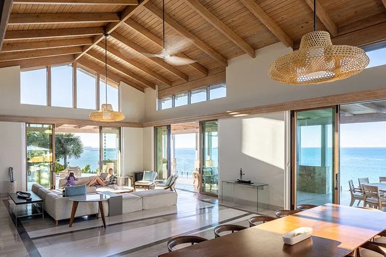 A spacious, modern living area with high wooden ceilings and large windows offering an ocean view at Oil Nut Bay. The room features a dining table, a seating area with couches, and hanging wicker lights. Glass doors open to a terrace that overlooks the sea.