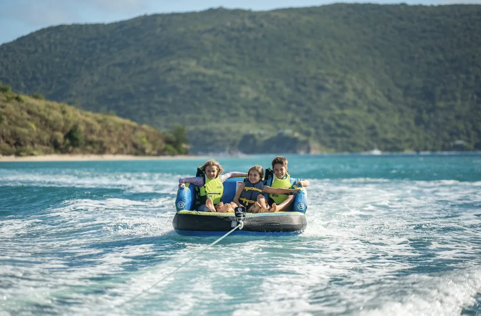 Three smiling children wearing neon yellow life vests enjoy a spirited tubing session on the vibrant, turquoise Caribbean Sea. In the background, the lush, green hills of Virgin Gorda meet a secluded sandy shoreline under a clear blue sky.