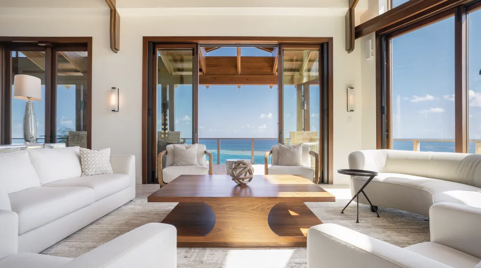 A bright, airy luxury villa living room features high vaulted wood ceilings and expansive floor-to-ceiling glass doors that open to a private deck. The bespoke interior includes plush white seating and a rich wood coffee table, framing a pristine view of the reef-protected bay and the horizon.