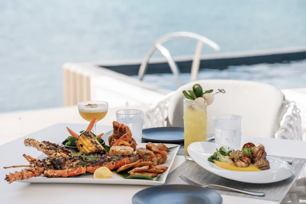 A vibrant close-up of a gourmet seafood platter featuring grilled Caribbean lobster, charred corn, and local sides, served poolside with two curated cocktails. In the background, the pristine blue water of the bay is visible past a sleek pool ladder.