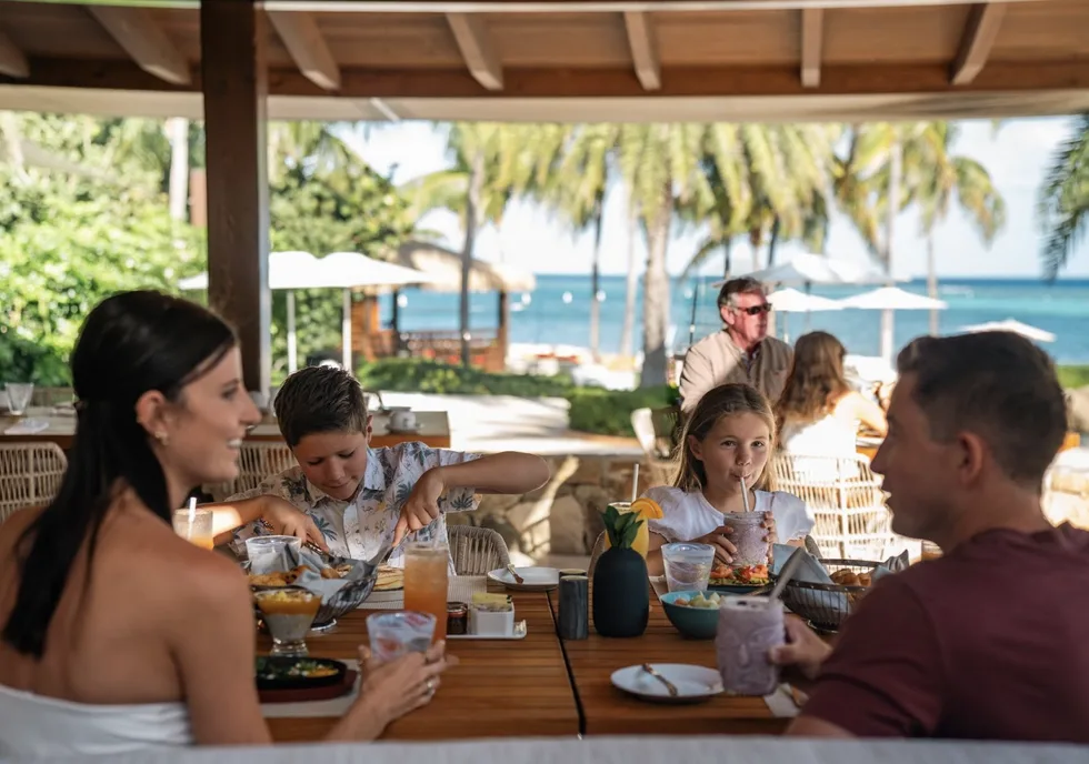 A family enjoys a relaxed, "barefoot luxury" breakfast at a long wooden table under a shaded outdoor pavilion. Beyond the diners, white umbrellas and palm trees line the secluded beach, meeting the turquoise reef-protected waters.