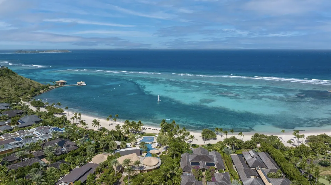 A spectacular aerial view of Oil Nut Bay's reef-protected waters, showcasing the vibrant transition from sandy-gold shallows to deep-blue sea. A lone white sailboat glides across the pristine bay, bordered by lush tropical hills and private luxury villas nestled along the secluded shoreline.