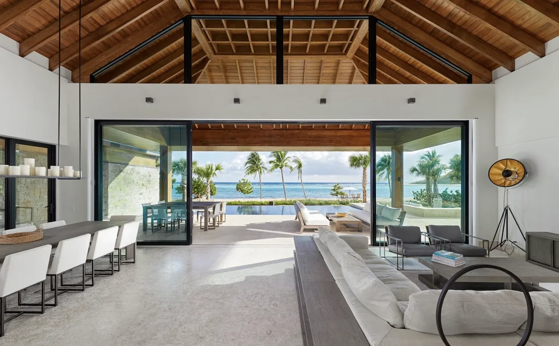 An expansive, contemporary living and dining area featuring high-vaulted wood ceilings and limestone flooring. Massive sliding glass doors offer a seamless transition to a private pool deck and beach, framing a curated view of palm trees and the turquoise Caribbean horizon.