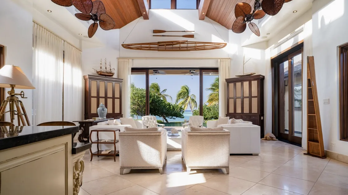 A sophisticated living room with a nautical theme, featuring a wooden rowing shell suspended from a vaulted ceiling and model ships on dark wood cabinets. Large glass doors open to a sun-drenched patio and pool, where the "Caribbean breeze" meets "barefoot luxury" interiors.