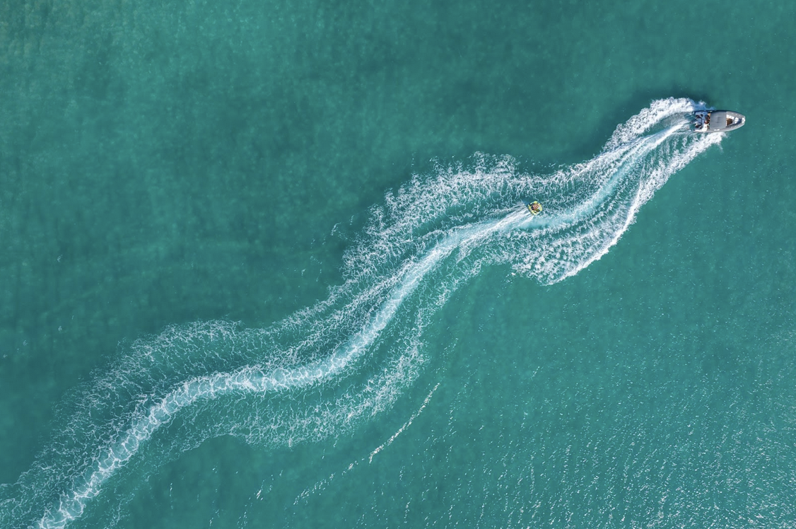 A spectacular aerial view of a motorboat carving a winding white wake through deep-blue Caribbean waters. A guest is being towed on a tube behind the boat, creating a vibrant scene of adventure and movement against the vast, reef-protected sea.
