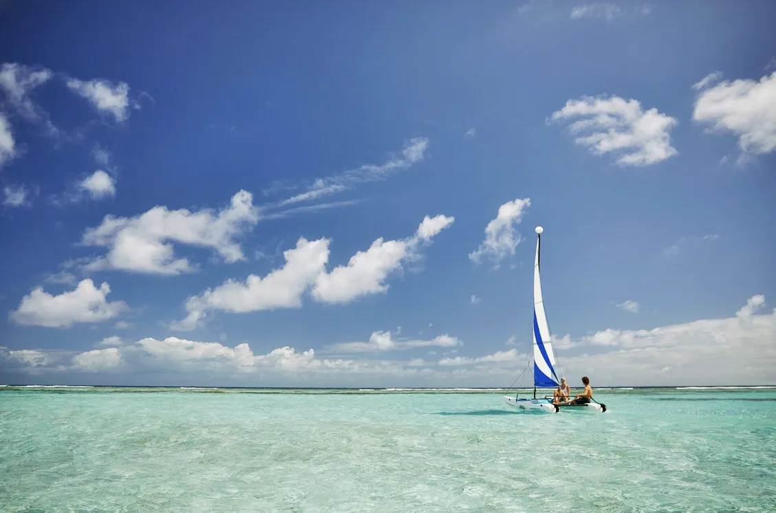 Two guests sail a white and blue Hobie Cat across the calm, pristine shallows of Oil Nut Bay. The crystal-clear water reveals the sandy-gold floor beneath, all under a bright blue sky filled with soft, white clouds.
