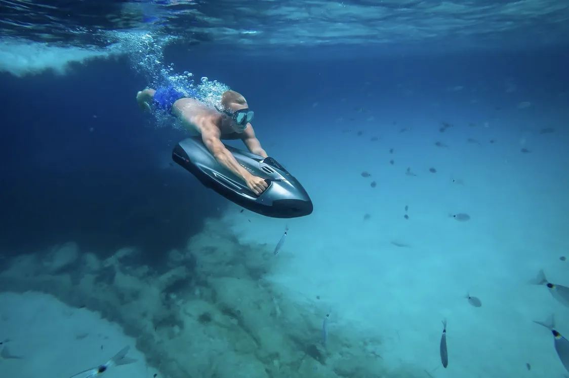 A guest wearing a mask and snorkel uses a sleek, grey underwater sea scooter to glide effortlessly through the deep blue sea. Small fish surround the diver as they explore the pristine aquatic world near the sandy ocean floor.