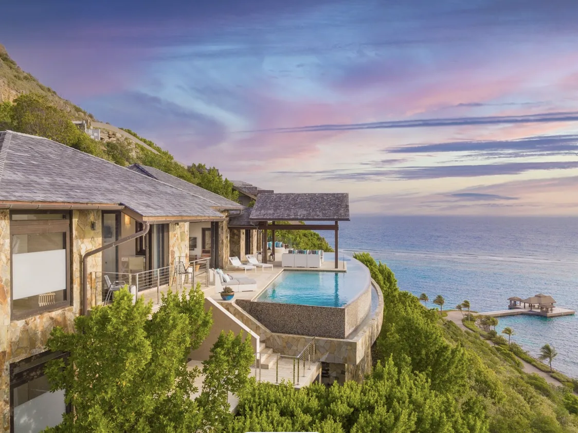 A spectacular twilight view of a multi-level luxury villa built into the lush green cliffs of Virgin Gorda. The stone-clad estate features a curved infinity pool that mirrors the purple and pink hues of the sunset sky, overlooking a private dock and the reef-protected bay below.