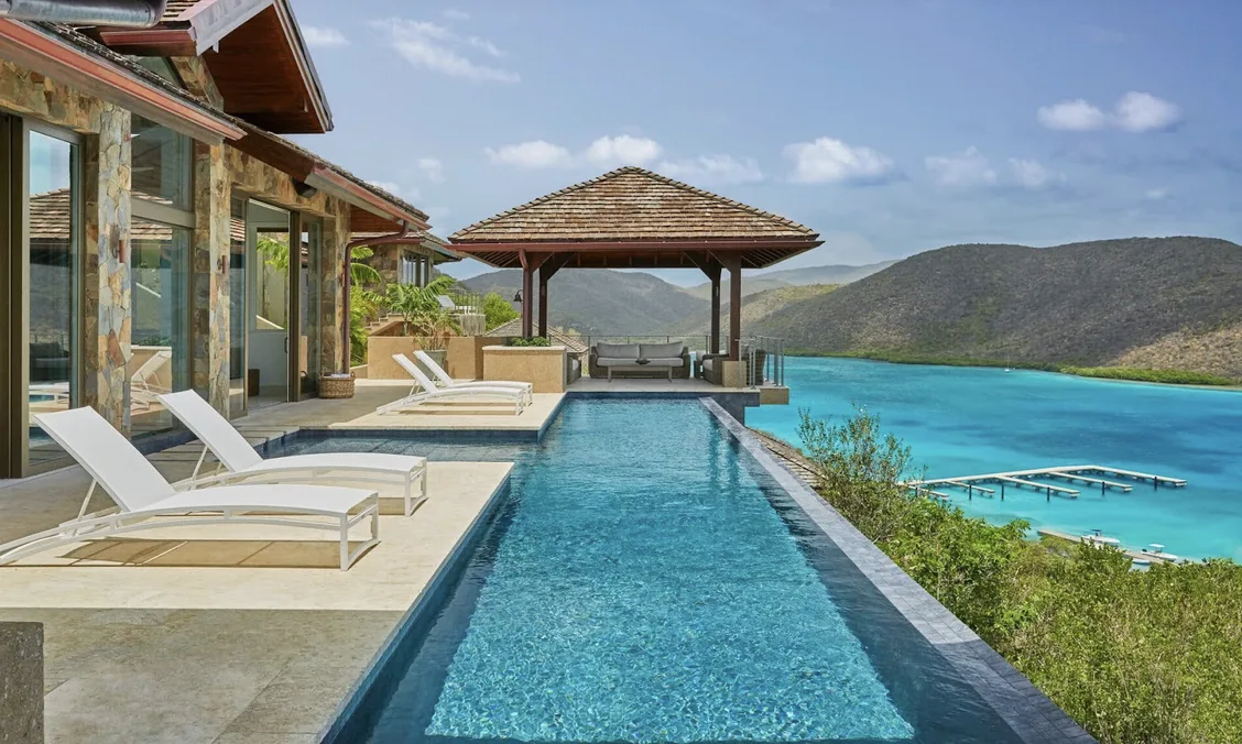 A private sanctuary featuring a long, turquoise infinity-edge pool that stretches toward the sea. White lounge chairs sit on a light stone patio next to a shaded wooden pavilion, offering a secluded vantage point of the lush island hills and the vibrant marina below.