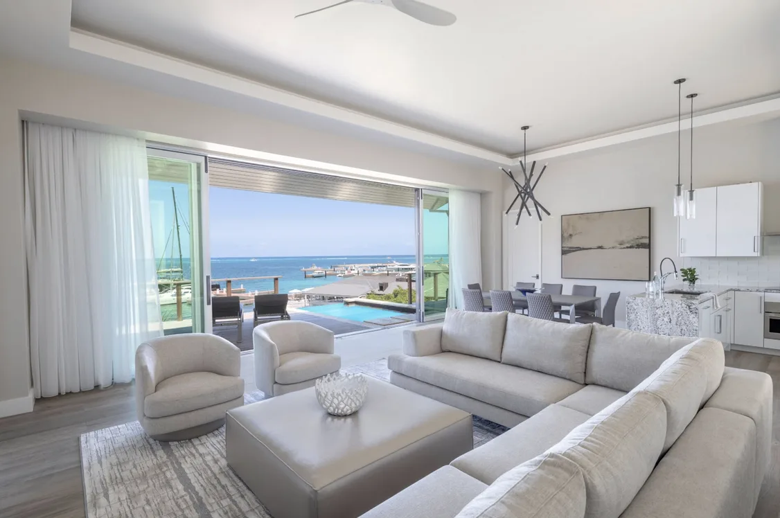 A modern, open-concept living area with a large grey sectional sofa and a sleek marble kitchen. The room opens completely to a private terrace and plunge pool, providing a curated view of the pristine bay and the white sailboats anchored in the distance.