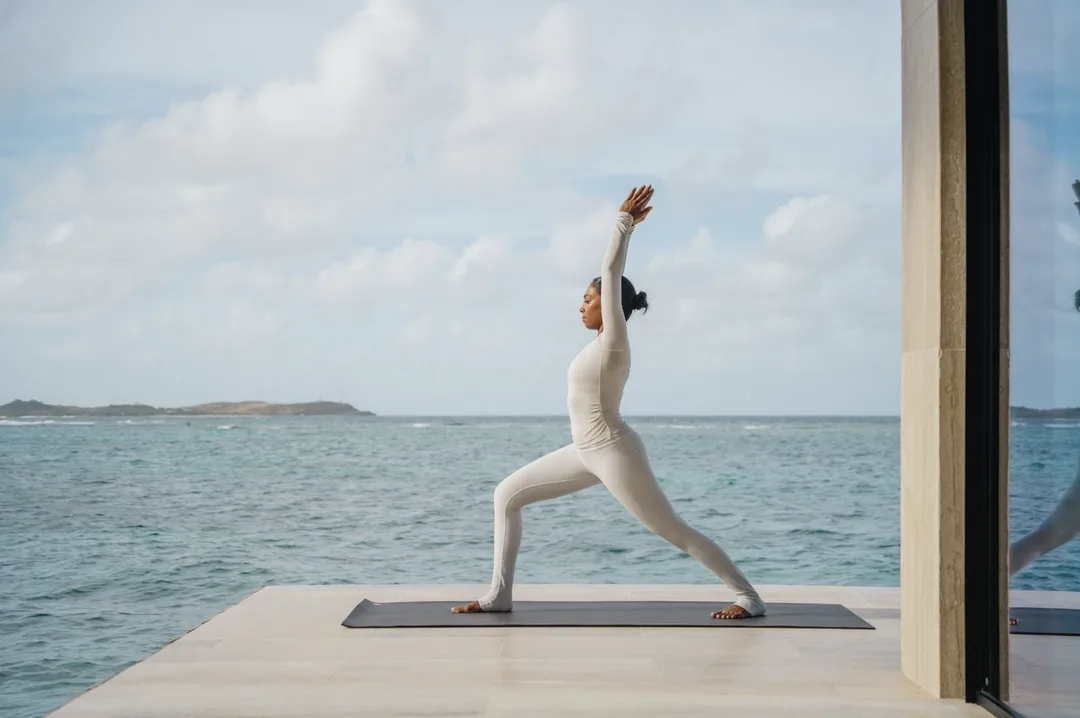 A woman in white athletic wear practices a yoga pose on a private stone deck extending over the reef-protected waters. The vast turquoise sea meets a soft horizon under a sky of scattered white clouds, embodying a quiet moment of "grounded and organic" luxury.