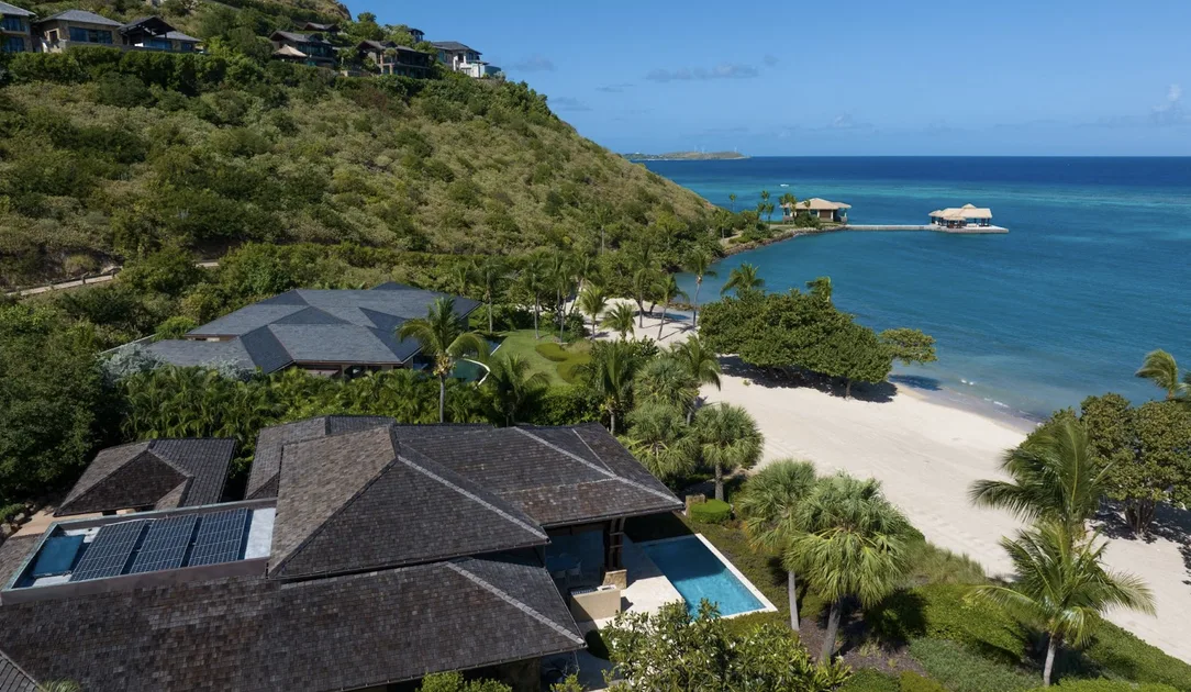 An elevated aerial view of a secluded luxury villa featuring dark-shingled roofs equipped with sleek solar panels. The estate is nestled into the lush green hills of Virgin Gorda, bordered by a pristine white-sand beach and the vibrant blue of the deep Caribbean Sea.