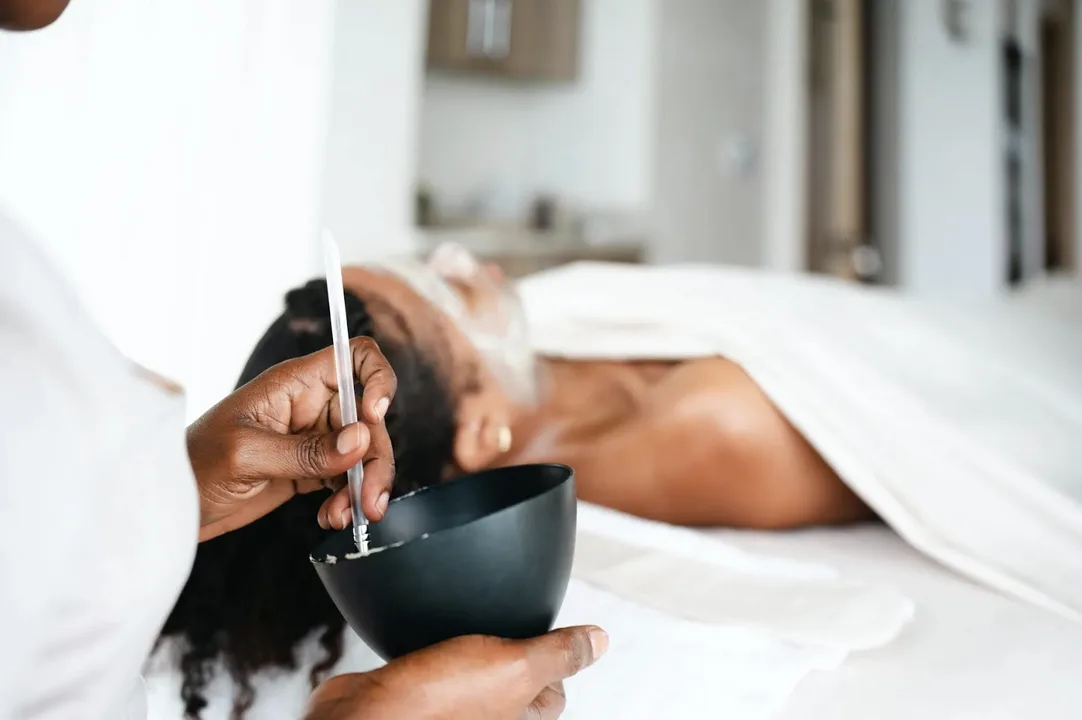 A close-up sensory shot of a spa professional's hands mixing a botanical treatment in a dark ceramic bowl. In the soft-focus background, a guest relaxes during a private treatment, illustrating the "concierge-level care" and "assured and effortless" service of the resort.