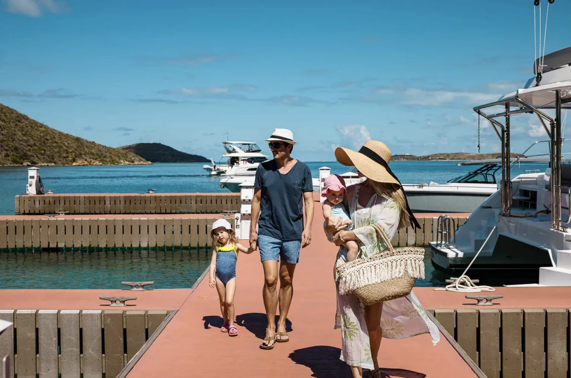 A family walks down a warm, sun-drenched wooden dock at the Oil Nut Bay marina. To their right, sleek white yachts are moored in the "vibrant" turquoise water, while the lush green hills of Virgin Gorda rise in the background under a clear "Deep Blue" sky.