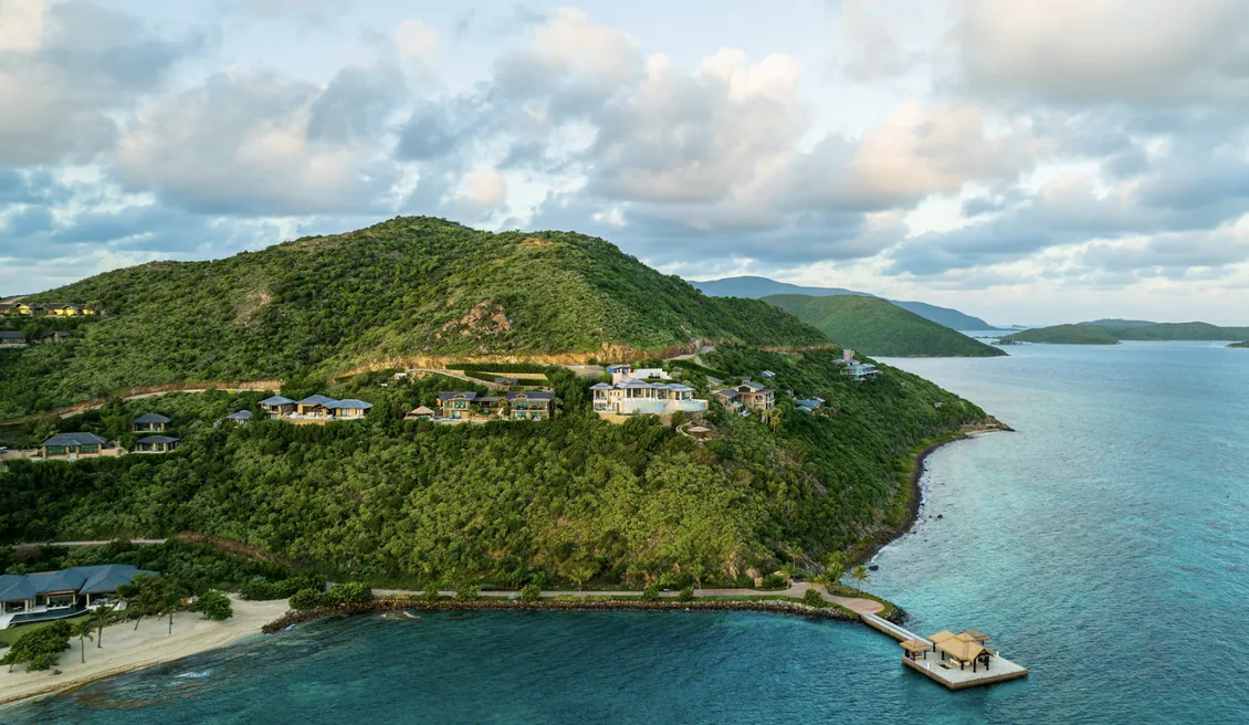A spectacular aerial view of the Oil Nut Bay peninsula, where luxury villas are "nestled into the cliffs" above a "secluded" white-sand beach. The "pristine" reef-protected bay surrounds a private arrival dock and overwater pavilion, showcasing the intersection of "untamed nature" and "uncompromised luxury".