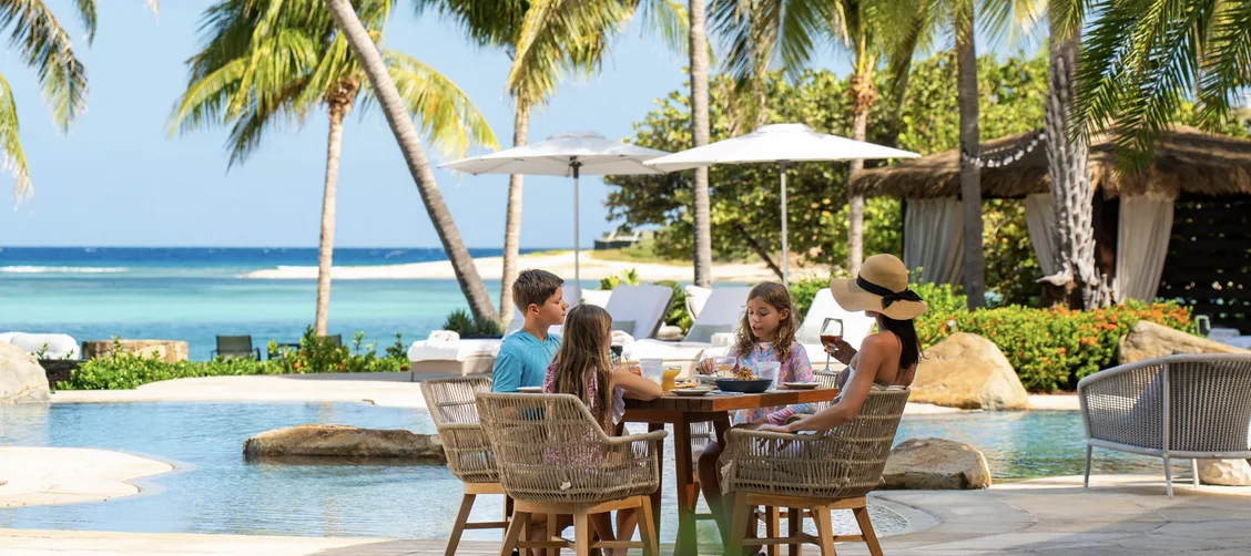 A family enjoys a "barefoot luxury" lunch at a wooden table beside a sparkling turquoise pool. White umbrellas and swaying palm trees provide shade, while a thatched-roof pavilion and the calm Caribbean Sea create a "spectacular" backdrop for "reconnecting with loved ones".