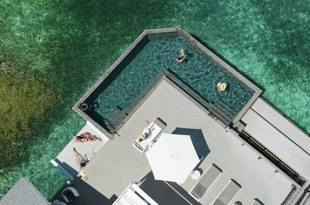 A spectacular aerial bird's-eye view of a modern overwater villa deck with a private, L-shaped infinity pool. Guests relax on white lounge chairs and swim in the "vibrant" turquoise water, which transitions into the "pristine" deep blue of the reef-protected bay.