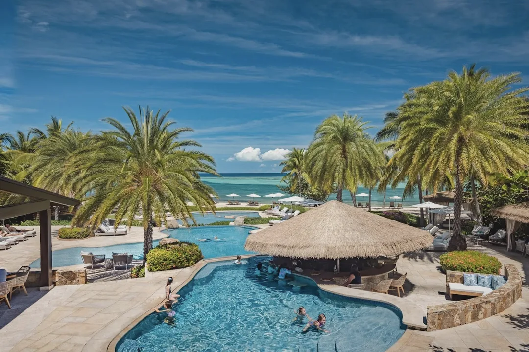 A vibrant scene at a luxury resort pool featuring a thatched-roof swim-up bar "nestled" among swaying palm trees. Guests enjoy the "pristine" multi-level pools while the reef-protected bay and a clear blue sky create an "intimate and exclusive" tropical backdrop.