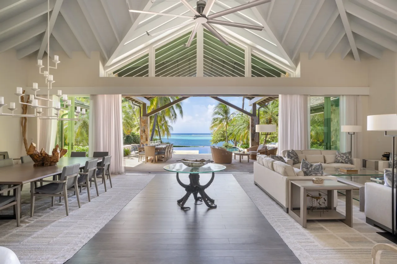 A bright, expansive great room in a luxury villa with high, vaulted ceilings and white exposed beams. Massive glass doors slide completely open to reveal a private patio, a turquoise infinity pool, and the deep blue horizon of the Caribbean Sea.
