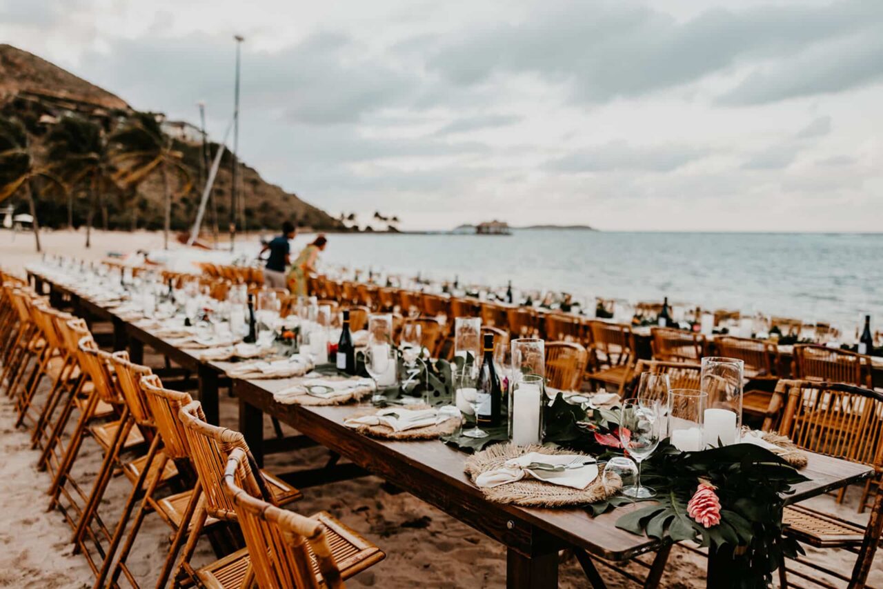 A long, bespoke wooden banquet table set for a luxury beach wedding at Oil Nut Bay, featuring organic floral arrangements, white candles, and elegant place settings on the pristine white sands of the British Virgin Islands.