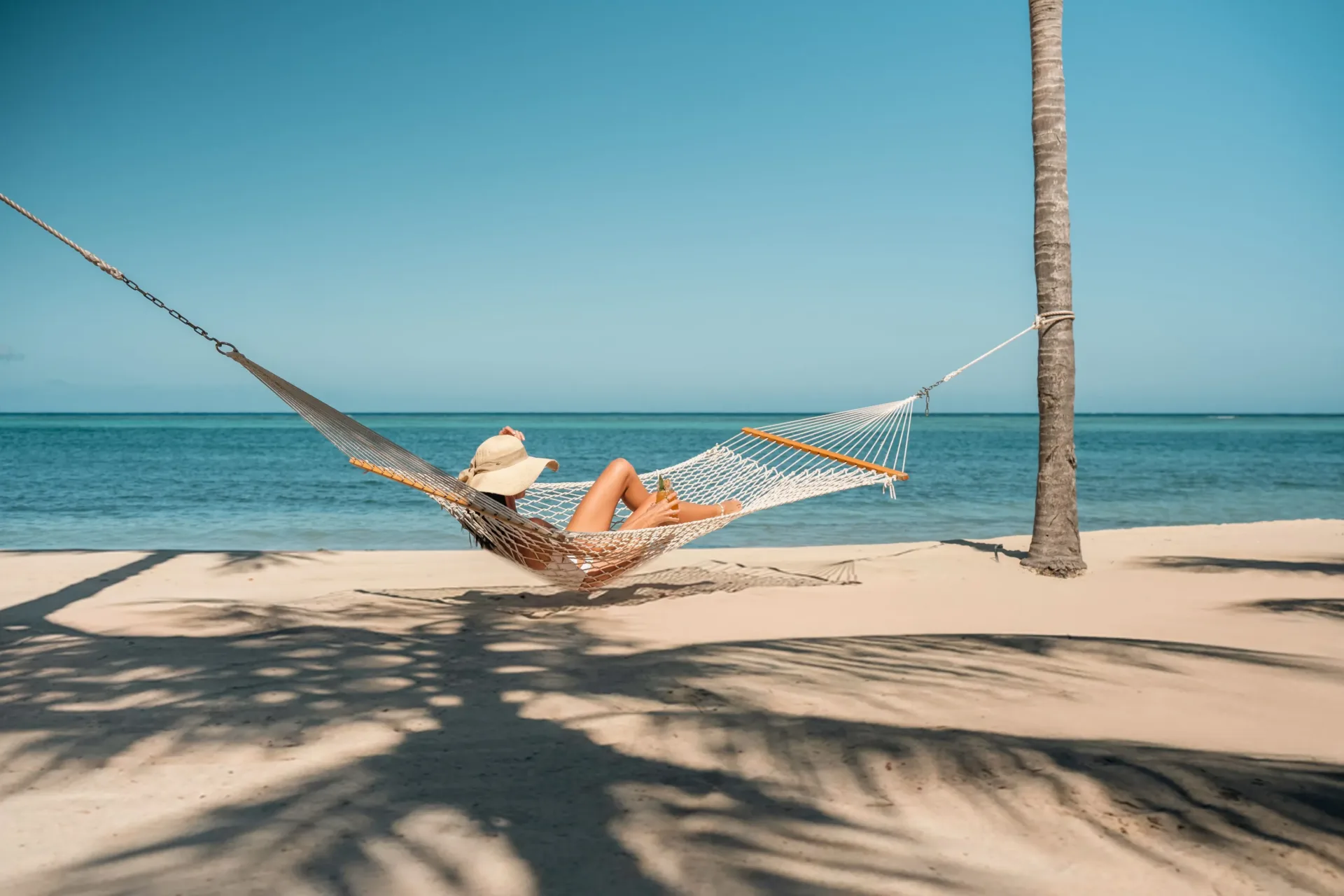 A serene moment of quiet luxury as a guest rests in a white woven hammock under a palm tree on a pristine, sandy gold beach beside calm Caribbean waters.