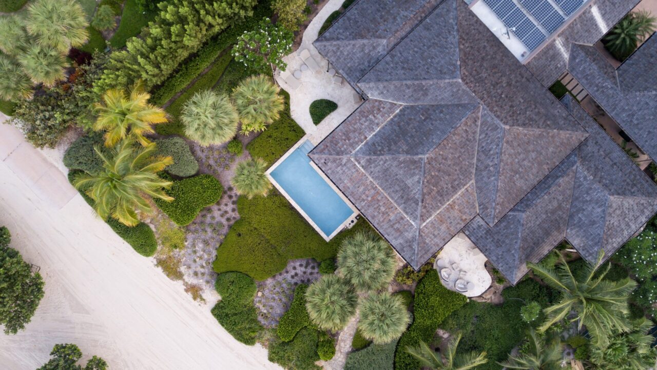 A top-down aerial view of a luxury estate showing dark-shingled rooftops nestled among lush tropical gardens and palm trees. A small, bright blue swimming pool is visible on the patio, and modern solar panels are integrated into one of the roof sections.