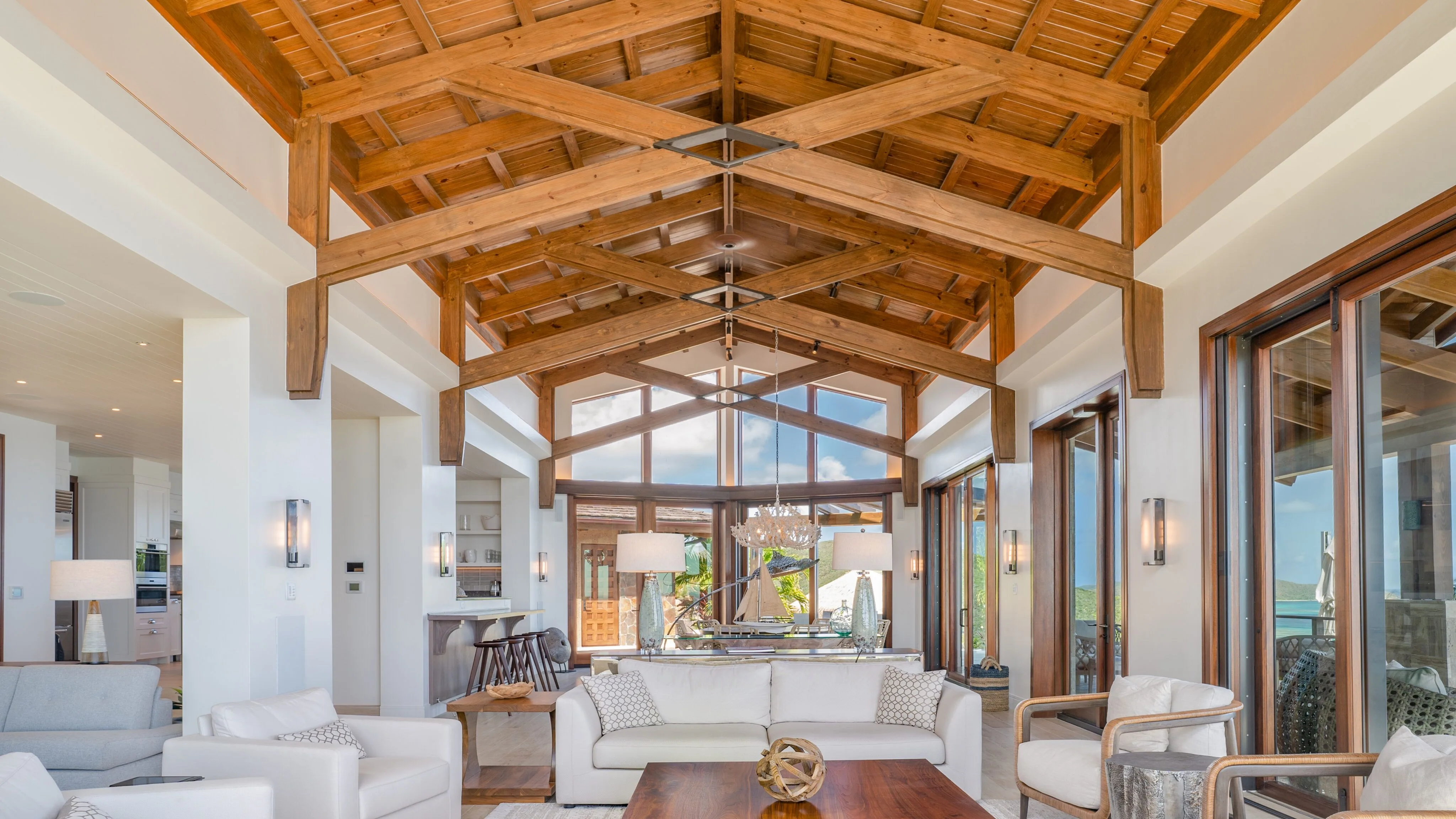 The interior great room of a luxury villa featuring high vaulted wooden ceilings, white modern furniture, and floor-to-ceiling glass doors that open to a sun-drenched Caribbean terrace.
