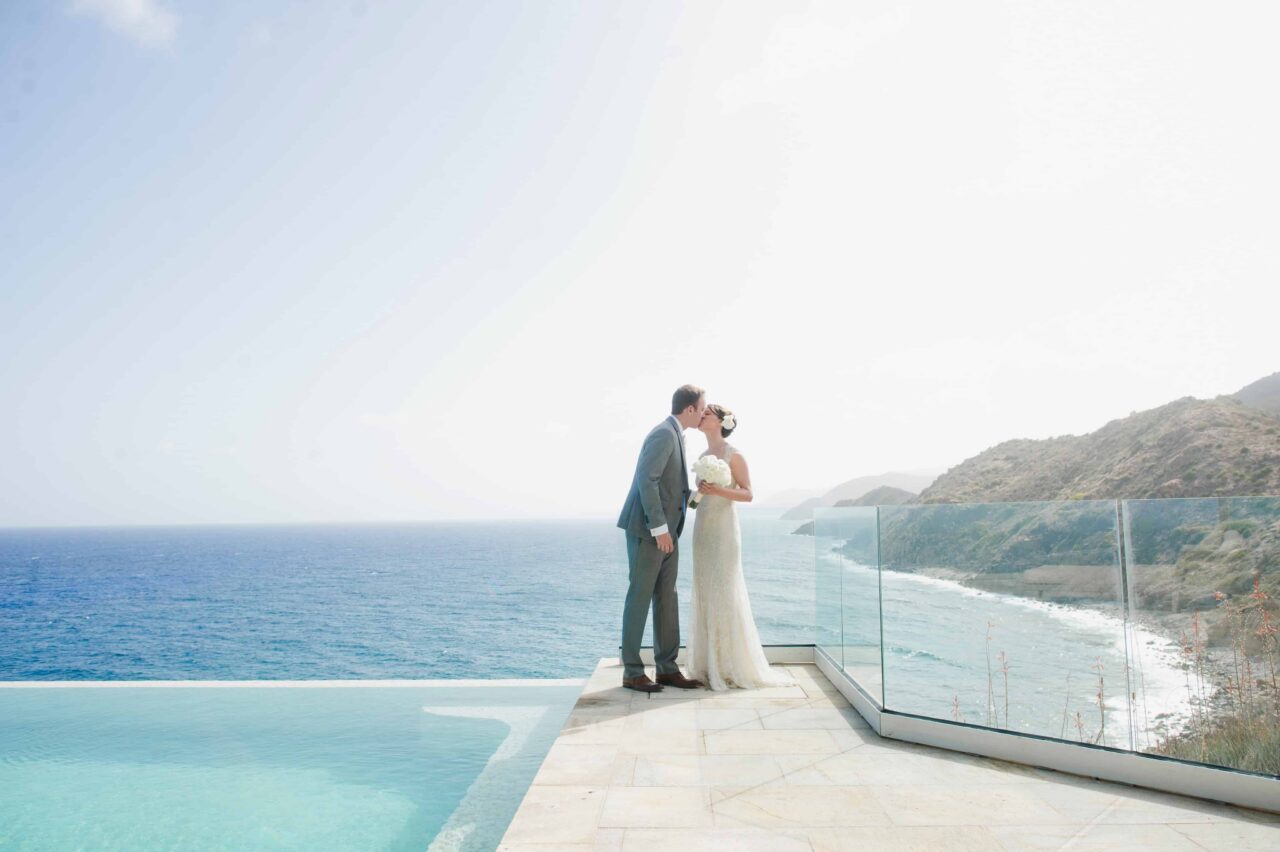 A bride and groom sharing a kiss on a modern stone terrace overlooking a dramatic coastline. The scene captures the intersection of uncompromised luxury and untamed nature, with steep cliffs and the deep blue sea stretching to the horizon.
