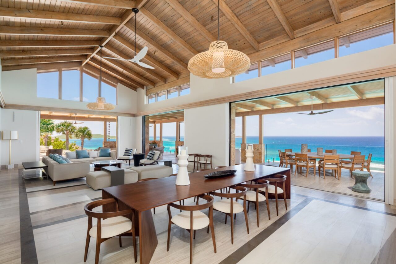 A wide-angle interior view of a contemporary villa with warm wood-slatted ceilings and neutral stone flooring. The open-concept space features a long, dark wood dining table and comfortable lounge seating, all overlooking a stunning, panoramic view of the turquoise ocean.