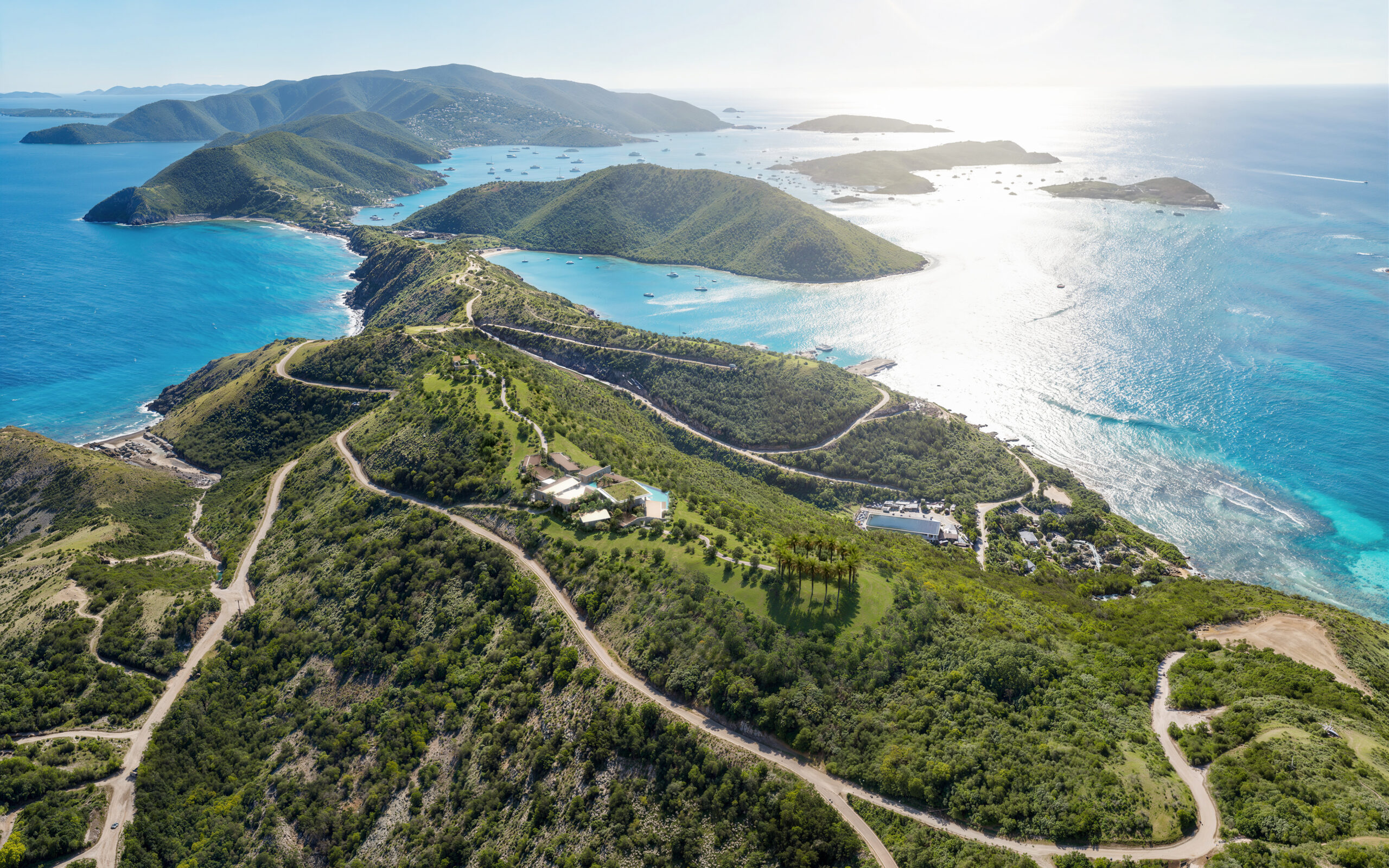 A sweeping, high-altitude aerial photograph of the lush, green Oil Nut Bay peninsula on Virgin Gorda. Winding roads lead to secluded luxury estates perched on the hillside, surrounded by vibrant reef-protected bays and the sparkling Atlantic Ocean.