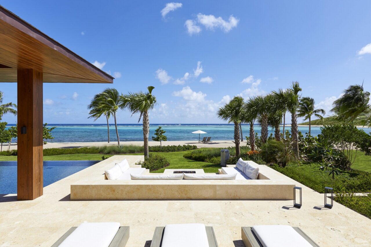 A luxury beachfront villa's outdoor lounge featuring a sunken stone fire pit area, a sparkling private infinity pool, and a view of the white sand beach and turquoise Caribbean Sea at sunset.