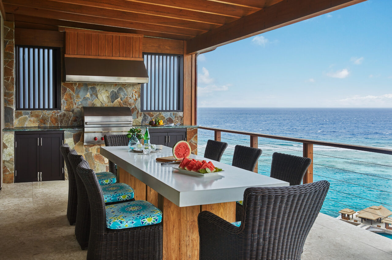 A wide outdoor dining terrace at a luxury villa with a long white table and modern chairs, overlooking a panoramic view of the deep blue Atlantic Ocean and Caribbean horizon.