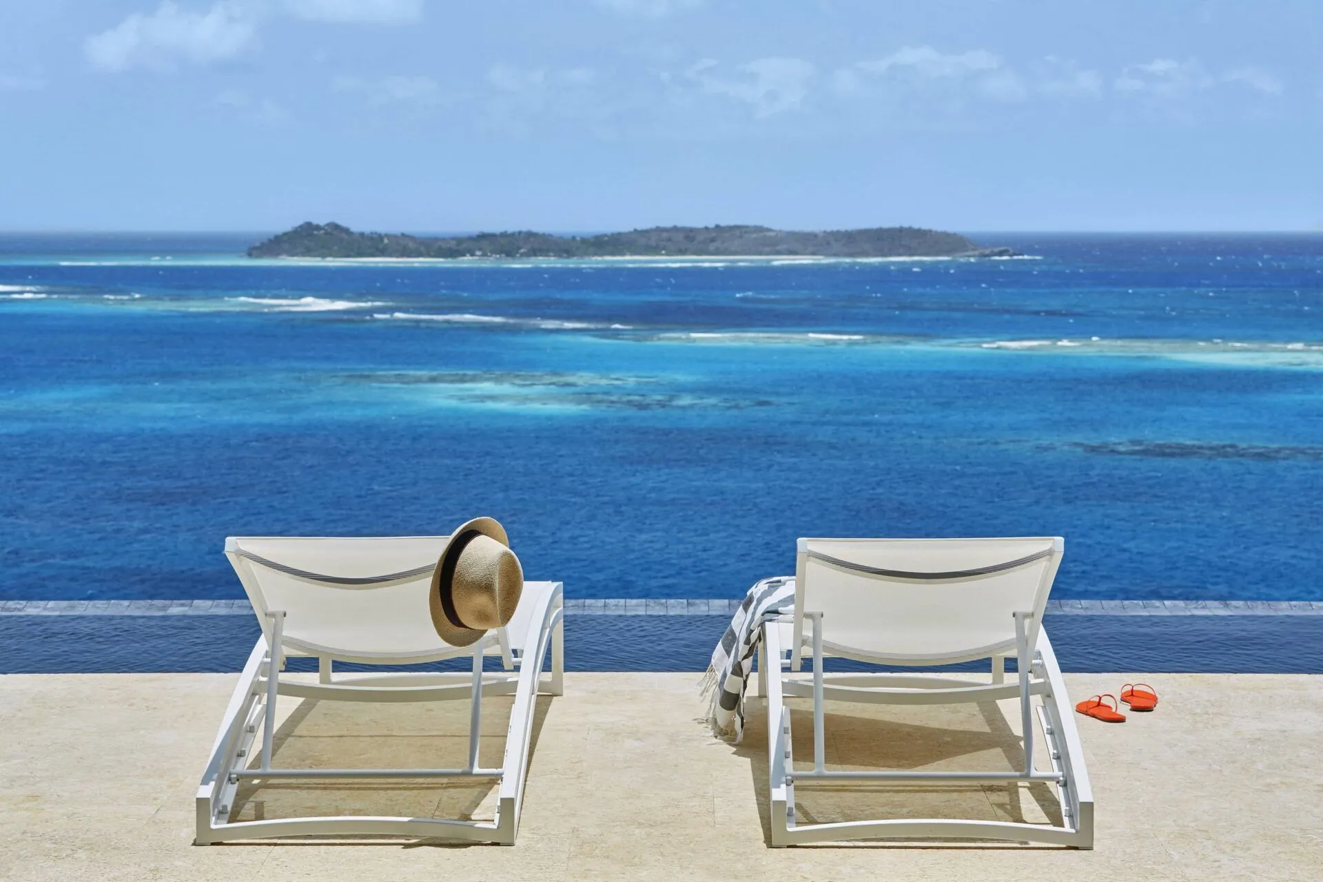 Two white lounge chairs set on a pristine sun deck beside a private infinity pool, offering an uninterrupted view of the deep blue Atlantic and a distant island.