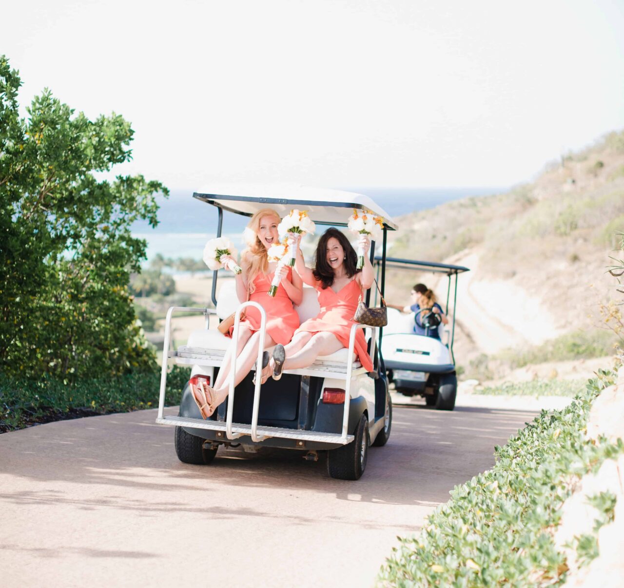 A joyful group of bridesmaids in coral dresses riding in the back of a white golf cart along a paved path. The background shows lush green hills and a distant view of the sea, highlighting the effortless and approachable island lifestyle.