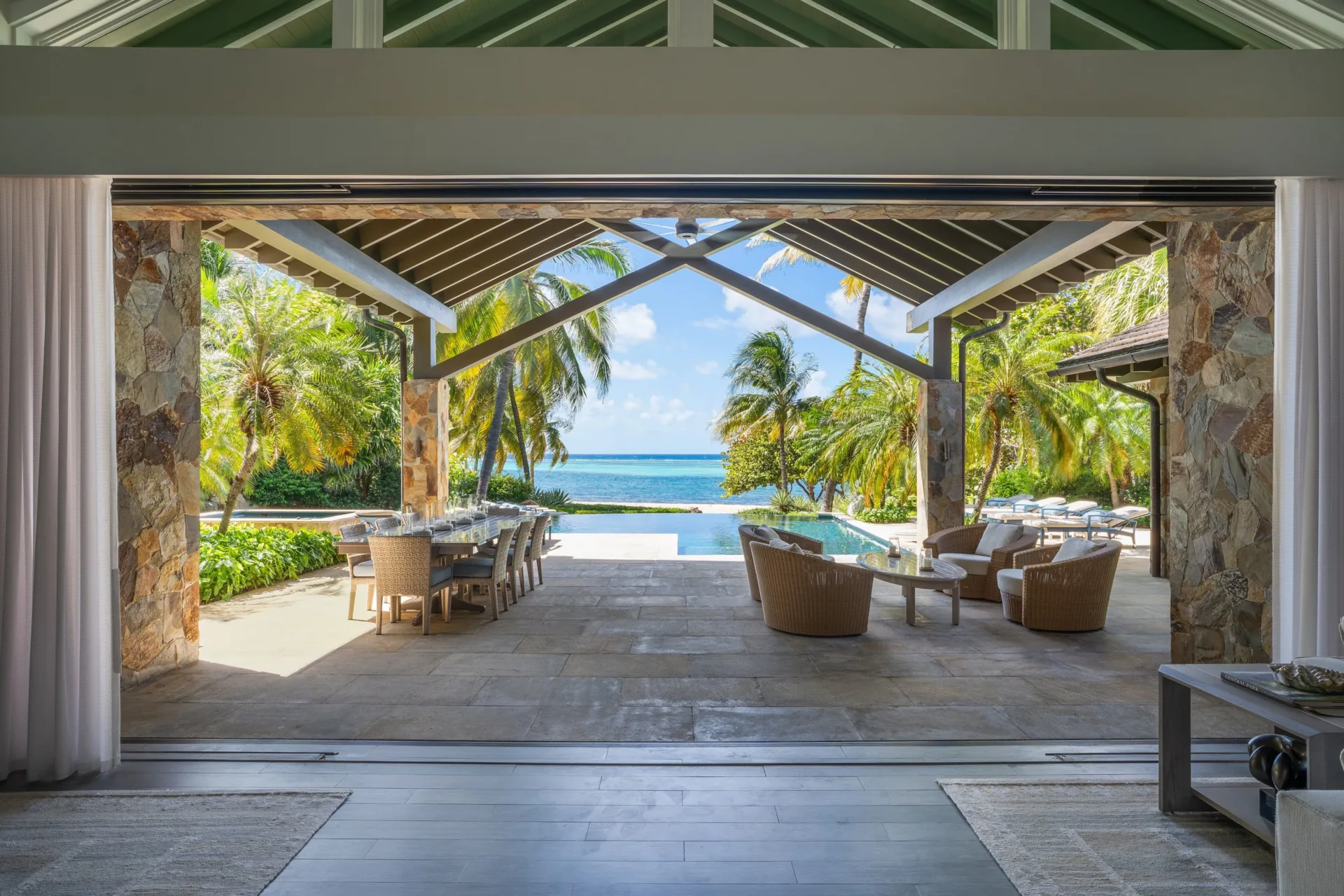 A wide view from a luxury villa's great room looking out onto a covered stone patio. The outdoor living space features an al fresco dining table and comfortable wicker seating next to an infinity pool, framed by lush palm trees and the turquoise waters of a reef-protected bay.