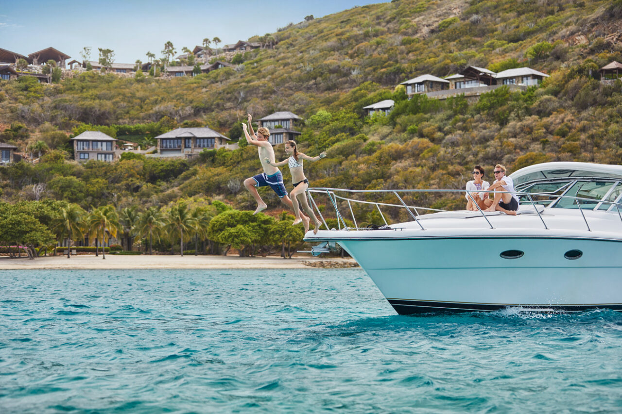 A bright, sunny day on the water where two teenagers are captured mid-air, jumping off the bow of a modern white yacht into clear turquoise water. On the boat's deck, an adult couple relaxes and watches them. In the background, a lush green hillside is dotted with luxury coastal villas overlooking a sandy beach with palm trees.