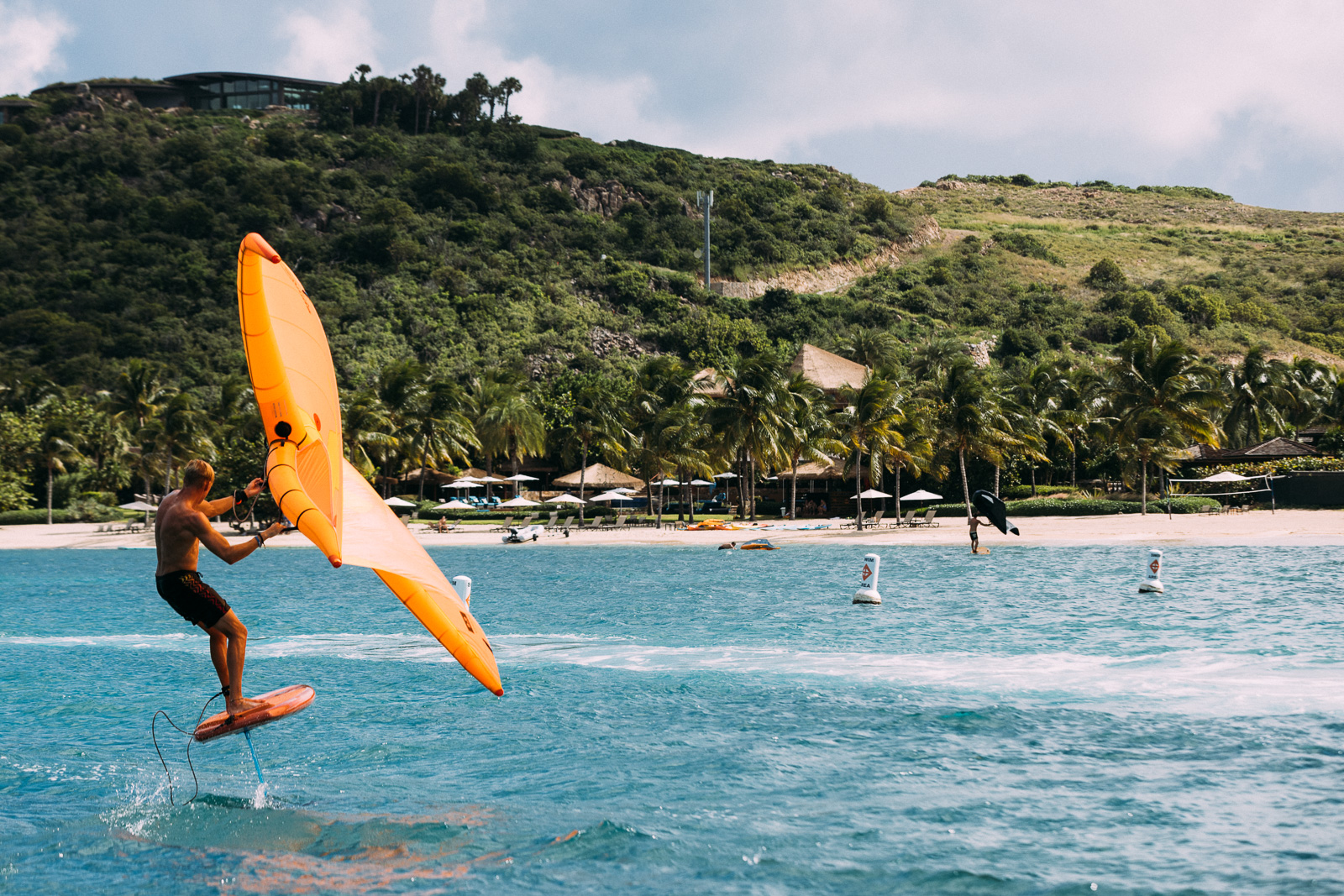 An action shot of a person wing foiling across vibrant blue water on a sunny day. The rider, wearing dark swim trunks, balances on a hydrofoil board that is lifted completely out of the water while holding a large, bright orange handheld wing sail. The background features a tropical beach lined with palm trees, white umbrellas, and a hillside with a modern resort structure.