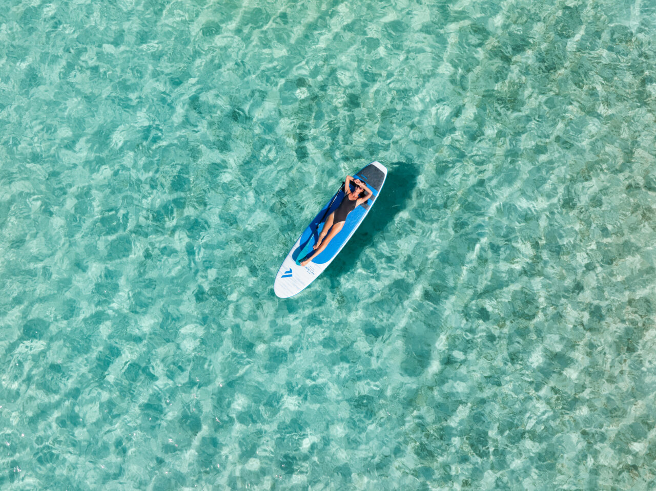 A high-angle, top-down aerial view of a woman in a black one-piece swimsuit relaxing on a blue and white stand-up paddleboard. She is lying on her back with one arm covering her eyes, floating peacefully in the middle of crystal-clear, shallow turquoise water that shows rippling patterns on the sandy ocean floor.